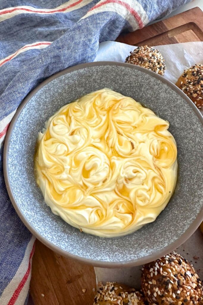 A grey stoneware dish of honey cream cheese dip that has had fresh honey swirled into the top using a skewer. You can see the sourdough discard bagel bites sitting around the sides of the bowl of dip.