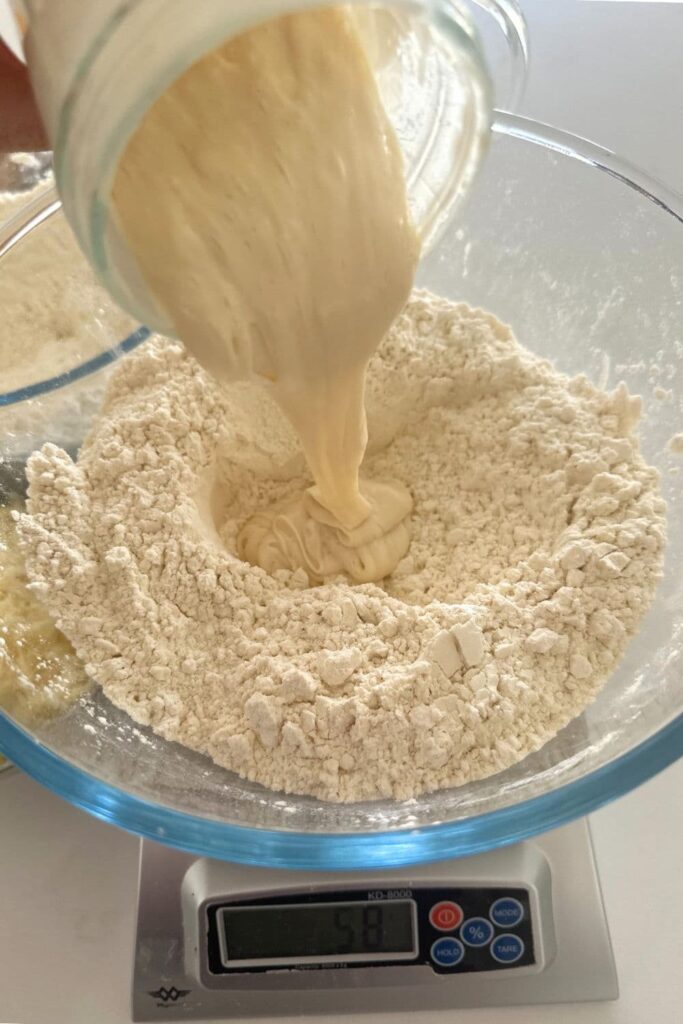 A bowl of flour that is sitting on a scale - there is a jar of sourdough starter being poured into the centre.