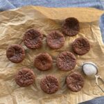 A selection of sourdough discard chocolate brownie bites sitting on a sheet of parchment paper. There is a small powdered sugar sifter and a blue dish towel in the photo also.