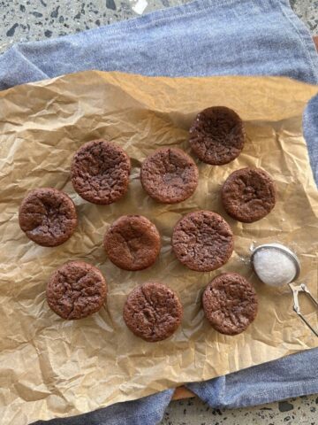 A selection of sourdough discard chocolate brownie bites sitting on a sheet of parchment paper. There is a small powdered sugar sifter and a blue dish towel in the photo also.