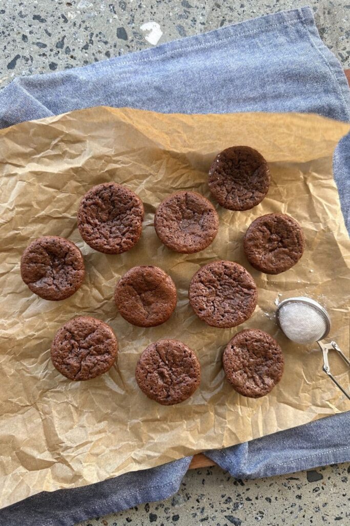 A selection of sourdough discard chocolate brownie bites sitting on a sheet of parchment paper. There is a small powdered sugar sifter and a blue dish towel in the photo also.
