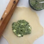 Sourdough gozleme dough rolled out with the spinach and feta filling sittin in the middle of the dough. There is a large rolling pin to the left of the sourdough gozleme dough.