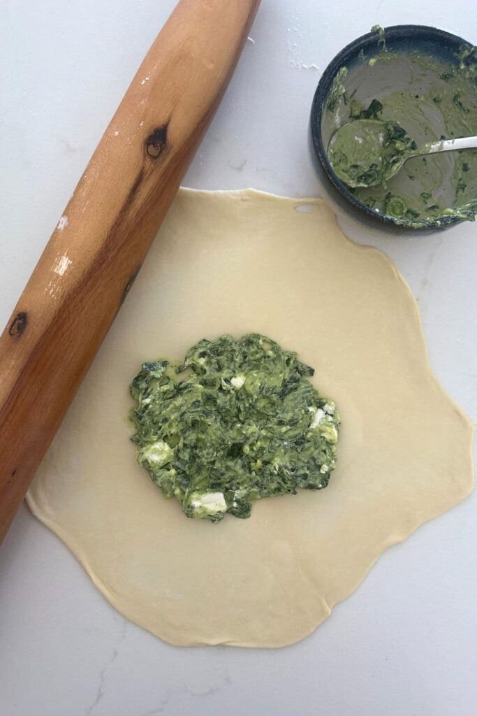 Sourdough gozleme dough rolled out with the spinach and feta filling sittin in the middle of the dough. There is a large rolling pin to the left of the sourdough gozleme dough.