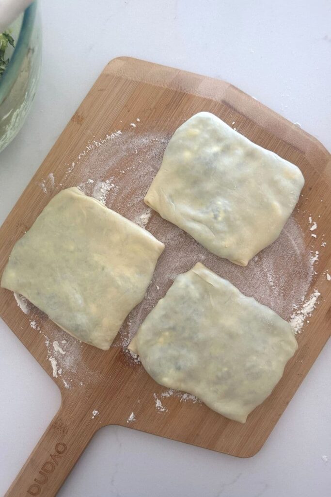 A wooden pizza peel dusted in flour. There are three uncooked sourdough gozleme sitting on top of the pizza peel. 