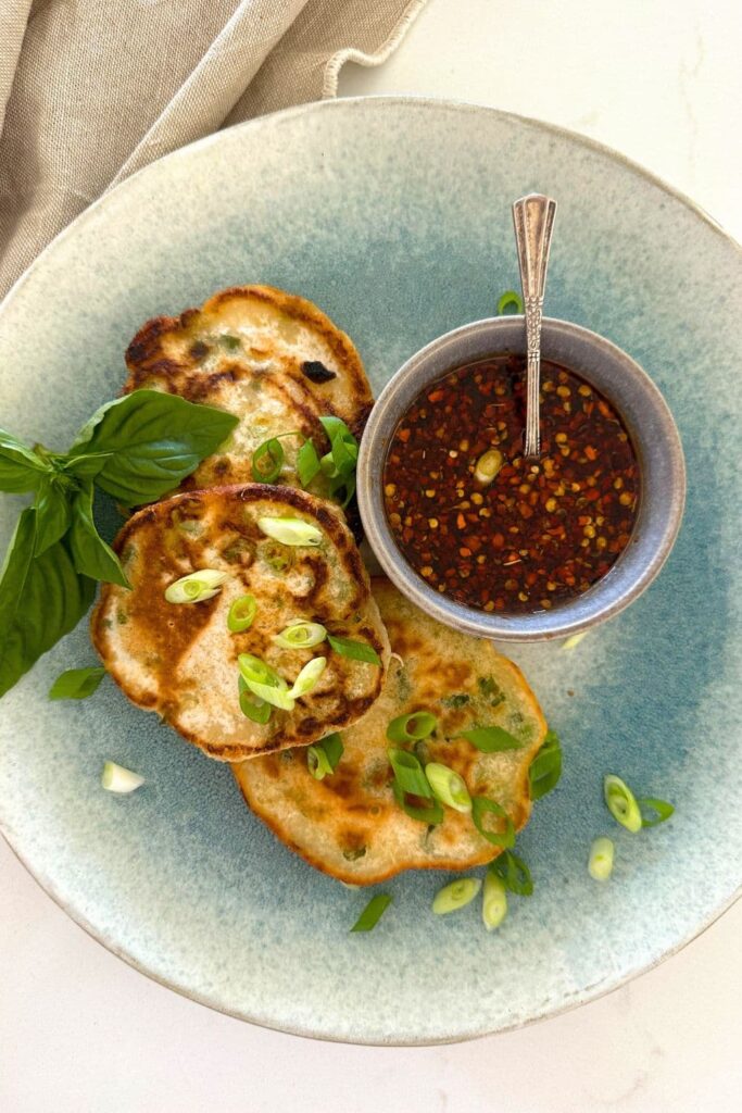 3 sourdough discard scallion pancakes sitting on a blue stoneware plate. There is also a bowl of chili soy dipping sauce sitting on the plate.