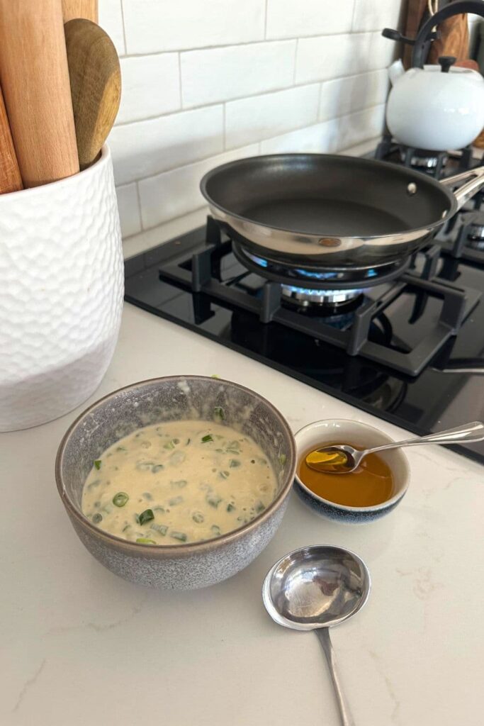A bowl of batter used to make sourdough discard scallion pancakes sitting on a white kitchen counter top next to a black cooktop.