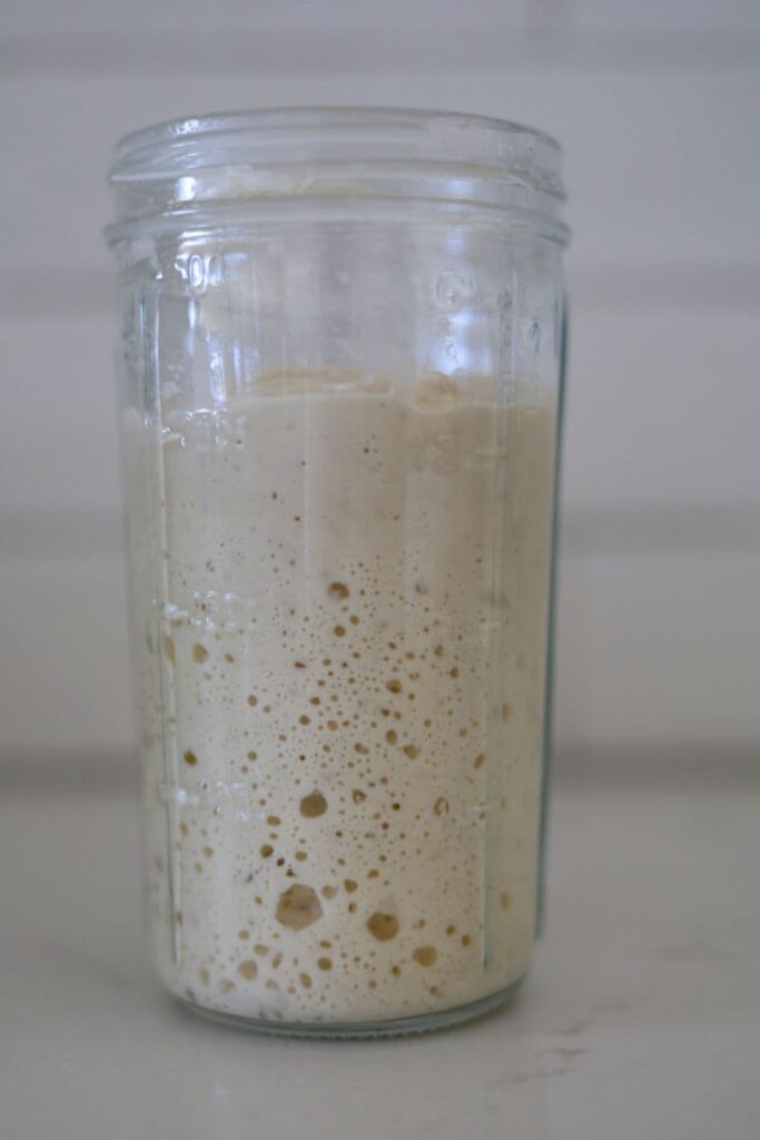 A jar of bubbly sourdough starter sitting on a white countertop.
