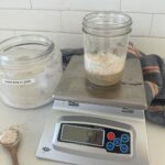 A photo of a 16oz mason jar sitting on a scale. The jar is filled with water and dark rye flour to create a rye flour sourdough starter. You can also see a large jar of rye flour in the background and a wooden spoon with rye flour on it next to the scale.