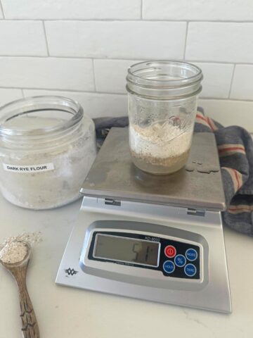 A photo of a 16oz mason jar sitting on a scale. The jar is filled with water and dark rye flour to create a rye flour sourdough starter. You can also see a large jar of rye flour in the background and a wooden spoon with rye flour on it next to the scale.