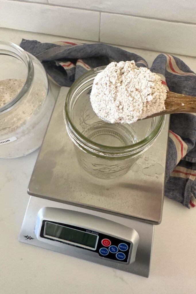 A wooden spoon filled with dark rye flour hovering over a 16oz mason jar sitting on a set of digital scales. The flour is being added to the jar of water to create a rye sourdough starter.