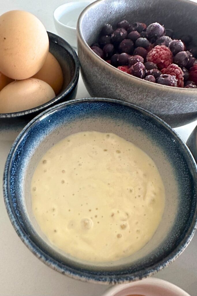 A bowl of sourdough starter sitting with a bowl of frozen berries and fresh eggs ready to be made into a sourdough clafoutis.