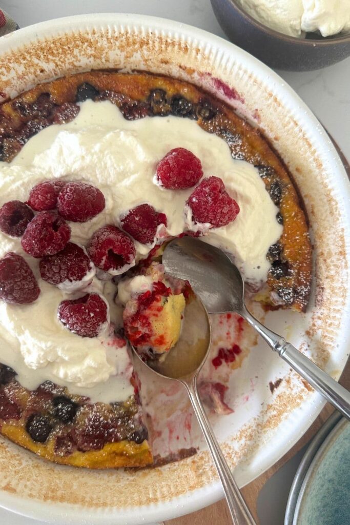 A sourdough mixed berry clafoutis that is topped with whipped cream and berries. A slice has been taken out and the spoon and slice are still sitting in the cream colored pie dish.