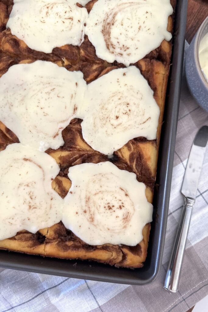 A sheet pan filled with sourdough cinnamon roll pancakes topped with vanilla frosting. You can see most of the pan. The right hand bottom corner of the photo is filled with a checked table napkin.