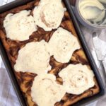 Sourdough cinnamon roll pancakes baked in a sheet pan and topped with vanilla icing. There is a bowl of icing to the side of the pancakes too. You can see the cinnamon swirls peaking from underneath the icing.