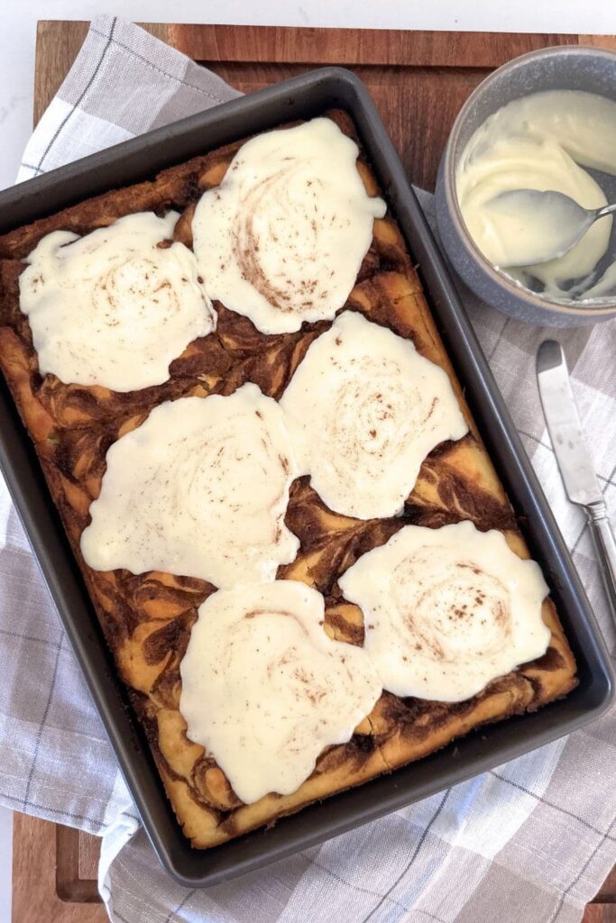 Sourdough cinnamon roll pancakes baked in a sheet pan and topped with vanilla icing. There is a bowl of icing to the side of the pancakes too. You can see the cinnamon swirls peaking from underneath the icing.