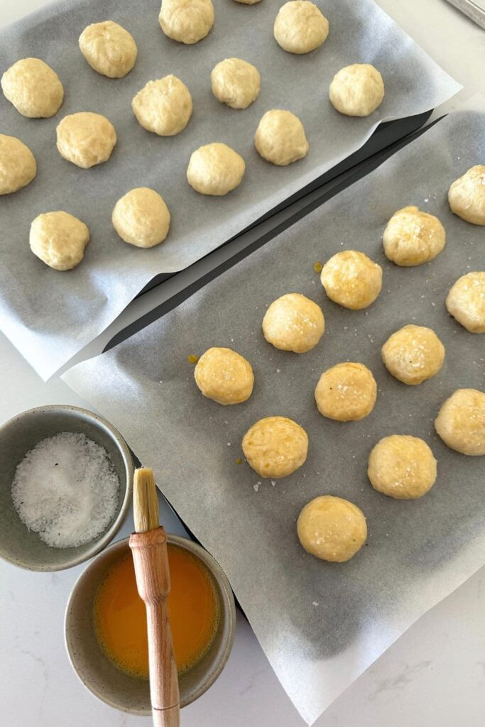 Sourdough discard pretzel bites that have been boiled and are sitting on a baking tray. They are being brushed with egg wash and sprinkled with coarse flaky sea salt.