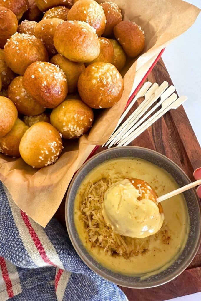 A tray of sourdough discard pretzel bites served next to a dish of 3 ingredient beer cheese dip. You can also see a blue and red dish towel in the photo. The whole display is sitting on a wooden board and there are a selection of wooden skewers in the photo too.