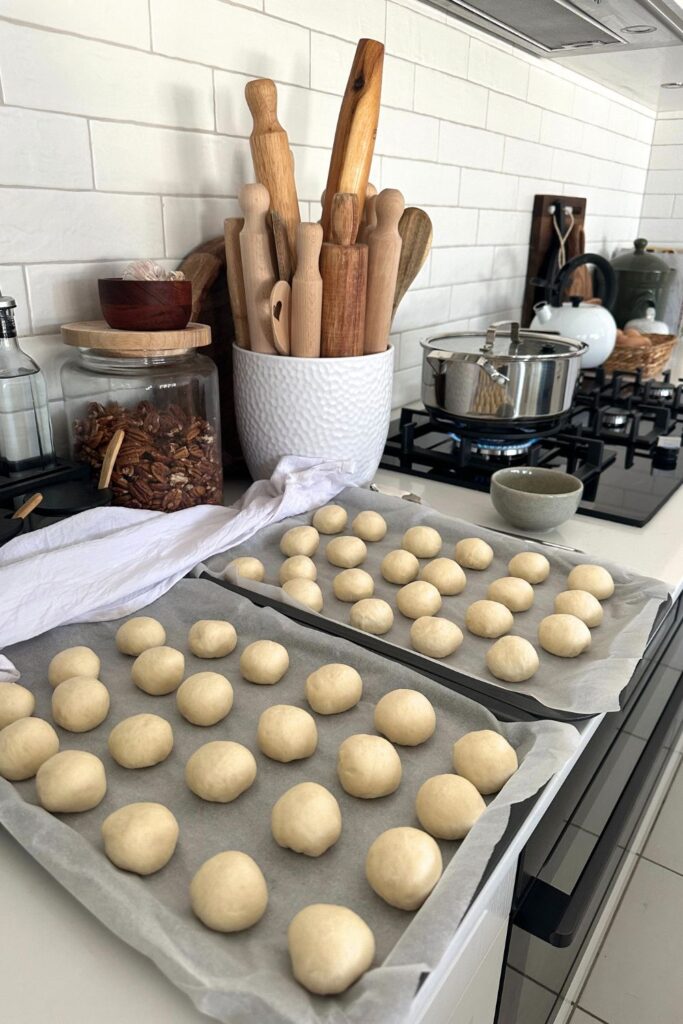 Sourdough discard pretzel bites sitting on a baking tray in a modern kitchen. You can see a pot of rolling pins in the background, as well as pot of water boiling on the stove top in preparation for boiling the sourdough discard pretzel bites.