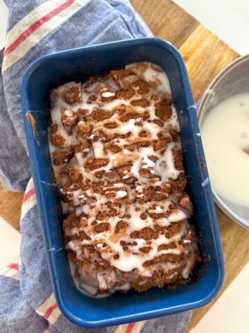 A loaf of sourdough discard apple fritter bread baked in a blue loaf pan. The sourdough apple fritter bread is being glazed with a vanilla glaze.