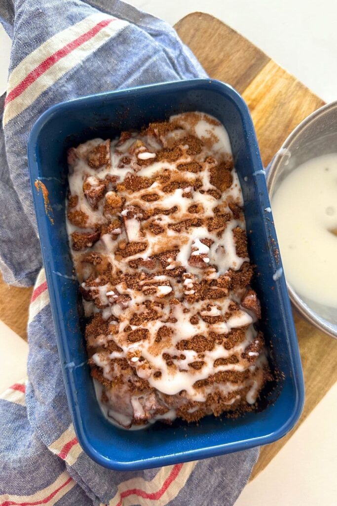 A loaf of sourdough discard apple fritter bread baked in a blue loaf pan. The sourdough apple fritter bread is being glazed with a vanilla glaze.