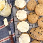 Sourdough blueberry cookies topped with lemon cream cheese frosting sitting on a wire cooling rack. To the left of the cookies you can see a blue and red striped dish towel and a jar of lemon cream cheese frosting with a wooden handled offset spatula.