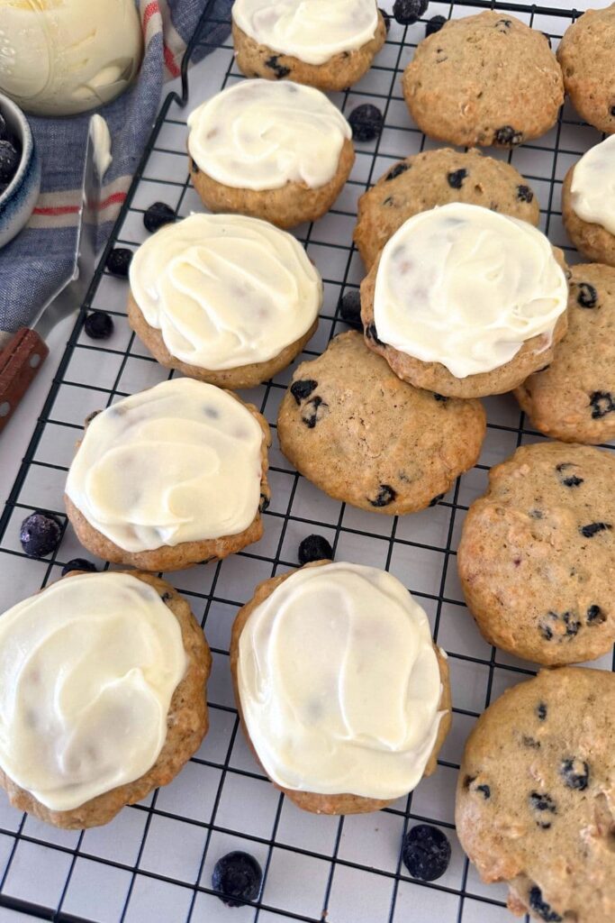An assortment of sourdough blueberry cookies sitting on a black wire cooling rack. Some of the cookies have lemon cream cheese frosting spread on top.