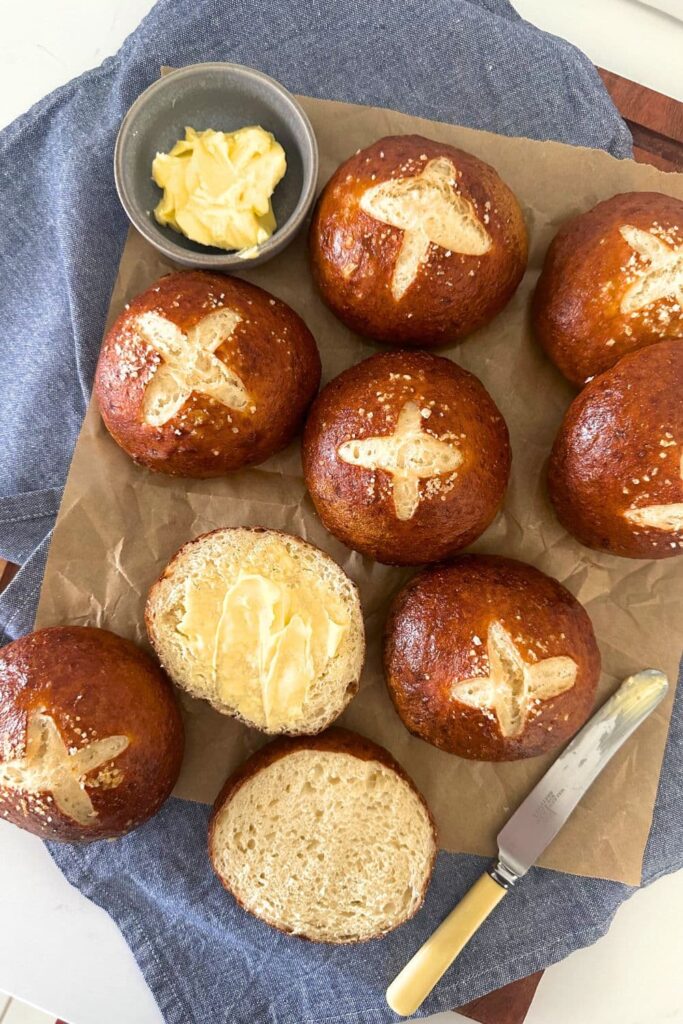 Sourdough pretzel buns sitting on a blue dish cloth. One has been sliced open to show the chewy crumb. There is an ivory handled bread knife and small pot of butter also in the photo.