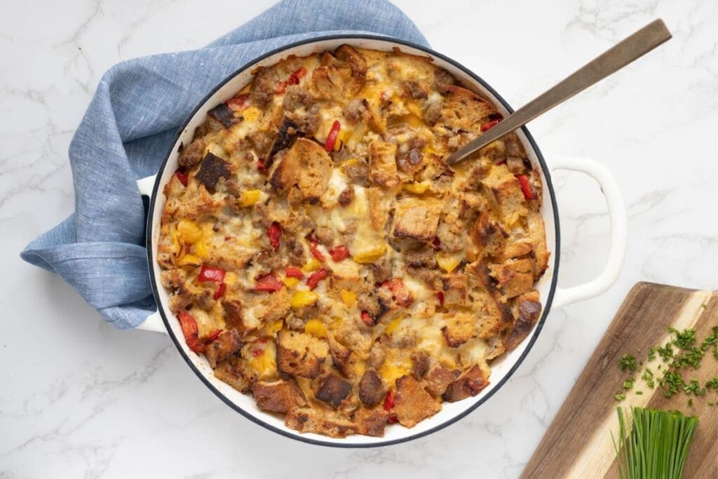 A round white cast iron skillet filled with a baked sourdough breakfast casserole. There is a wooden board with some chopped fresh chives in the bottom right hand corner.