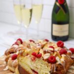 A sourdough brioche braided loaf decorated with flaked almonds and fresh raspberries displayed on a wooden board. There are 3 glasses of champagne and a bottle of Moet in the background of the photo.