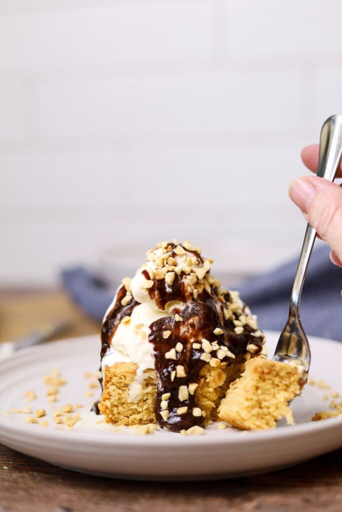 A sourdough peanut butter square on a white plate topped with vanilla ice cream, chocolate sauce and crushed peanuts. There is a fork taking a piece of the peanut butter square.