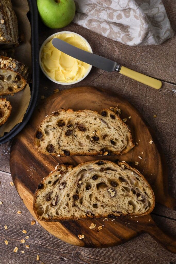 2 slices of sourdough apple crisp bread laid on a round wooden board so you can see the crumb inside. Around the slices of bread you can see a dish of butter, a floral dish towel and a green apple.
