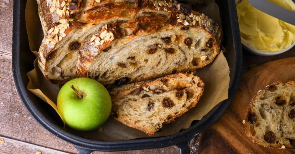 Sourdough apple crisp loaf sliced open so you can see the inside. The loaf is displayed inside a cast iron bread pan with a green apple next to it.