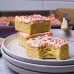 A slice of sourdough sugar cookie bar topped with strawberry frosting and rainbow sprinkles displayed on a white plate. There is a bite taken out of the slice and two other pieces behind it stacked on top of each other.