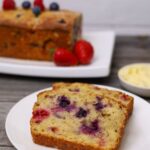 A feature image of sourdough berry banana bread. There are two slices of sourdough berry banana bread displayed on a white plate. You can see the rest of the loaf in the background of the photo garnished with fresh berries.