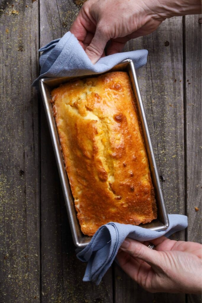 A sourdough cornbread loaf baked in a silver loaf pan. The pan is being held by a pair of hands with blue napkins.