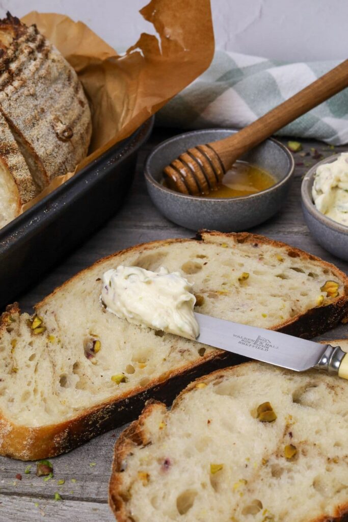 A photo of 2 slices of sourdough pistachio bread that has been displayed on a wooden board with a dish of cream cheese and a dish of honey. There is a knife with cream cheese laid on the slices of bread. You can see the rest of the loaf in the background of the photo.