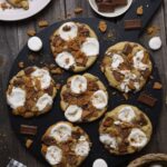 6 sourdough s'mores cookies sitting on a round black serving board. There are some decorative items surrounding the board, but these are ingredients that have been used in the sourdough s'mores cookies.