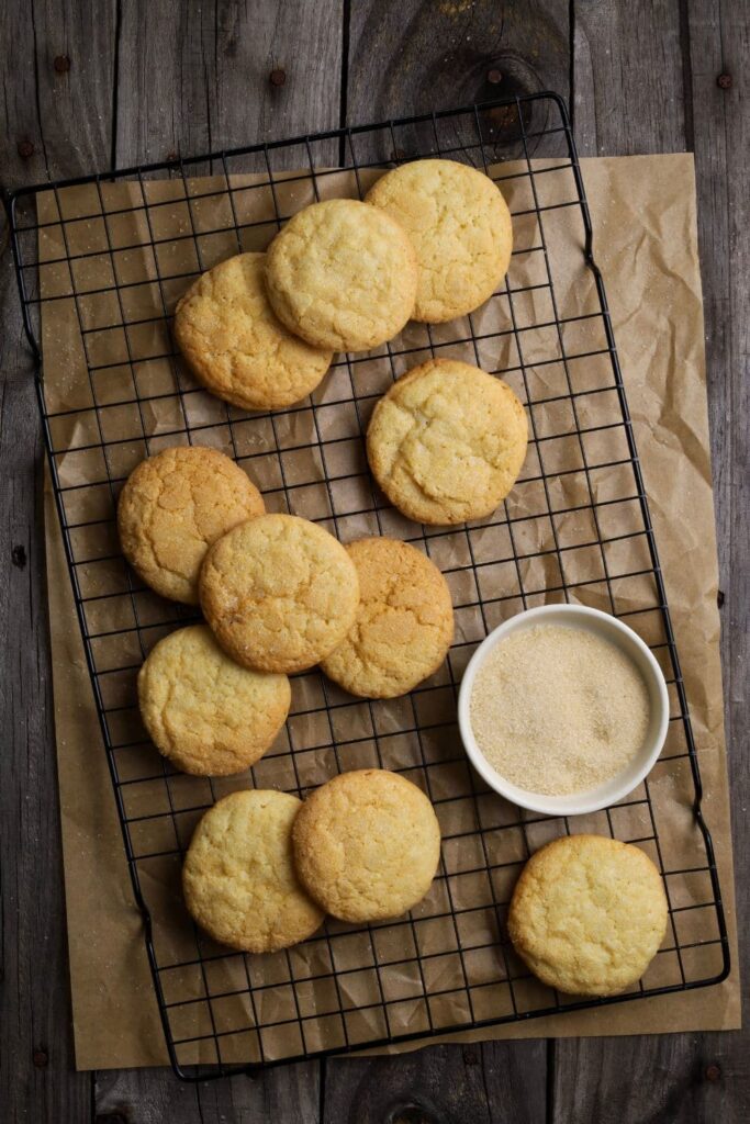 Sourdough vanilla wafers laid out on a black wire cooling rack. There is a piece of parchment paper under the wire cooling rack and a small dish of granulated sugar on the tray with the cookies.