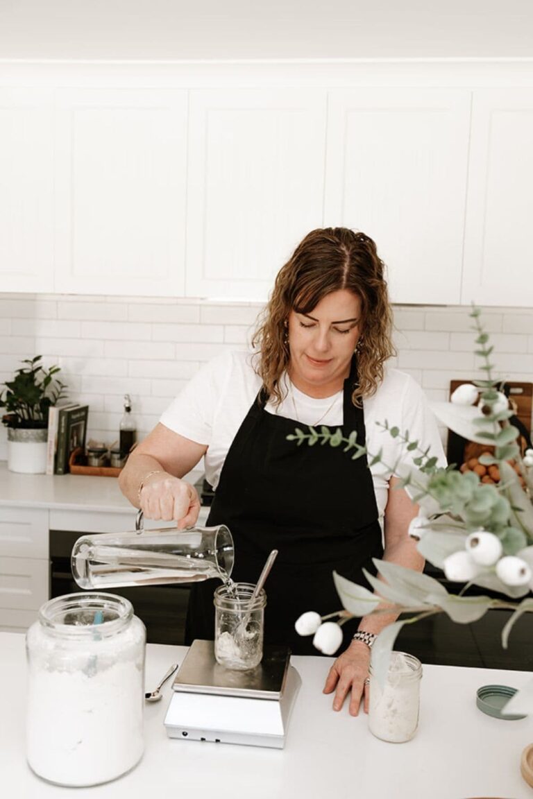 Kate from The Pantry Mama in the kitchen feeding her sourdough starter at the kitchen counter. She is using a kitchen scale to measure water from a jug into a mason jar. There is a jar of flour on the counter in front of her.