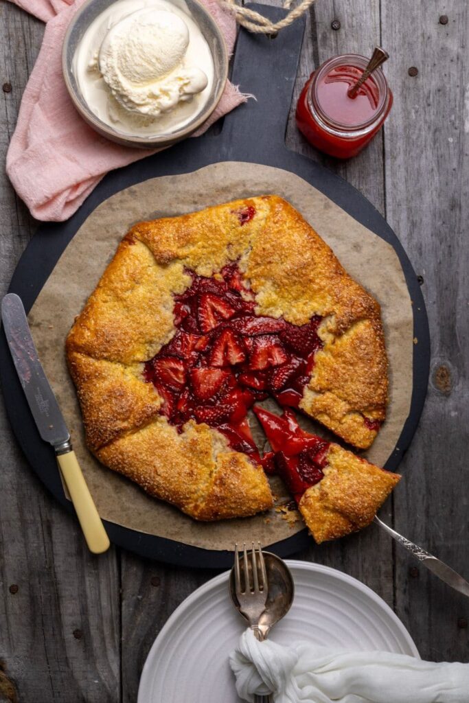 A vertical photo of a sourdough discard strawberry galette brimming with red strawberries. A slice has been taken out of the galette, but is still sitting on the plate. There is a bowl of vanilla ice cream sitting on a pink linen napkin above the galette and a small jar of strawberry sauce.