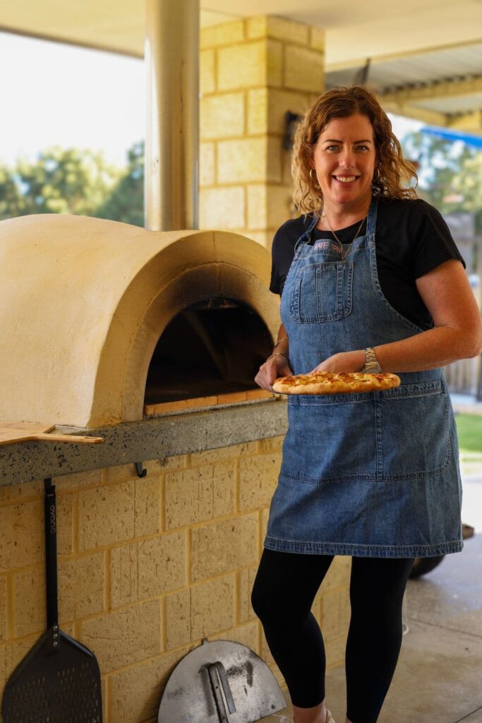 Kate from The Pantry Mama standing in front of a wood fired pizza oven with a sourdough pizza that she just baked in front of her.