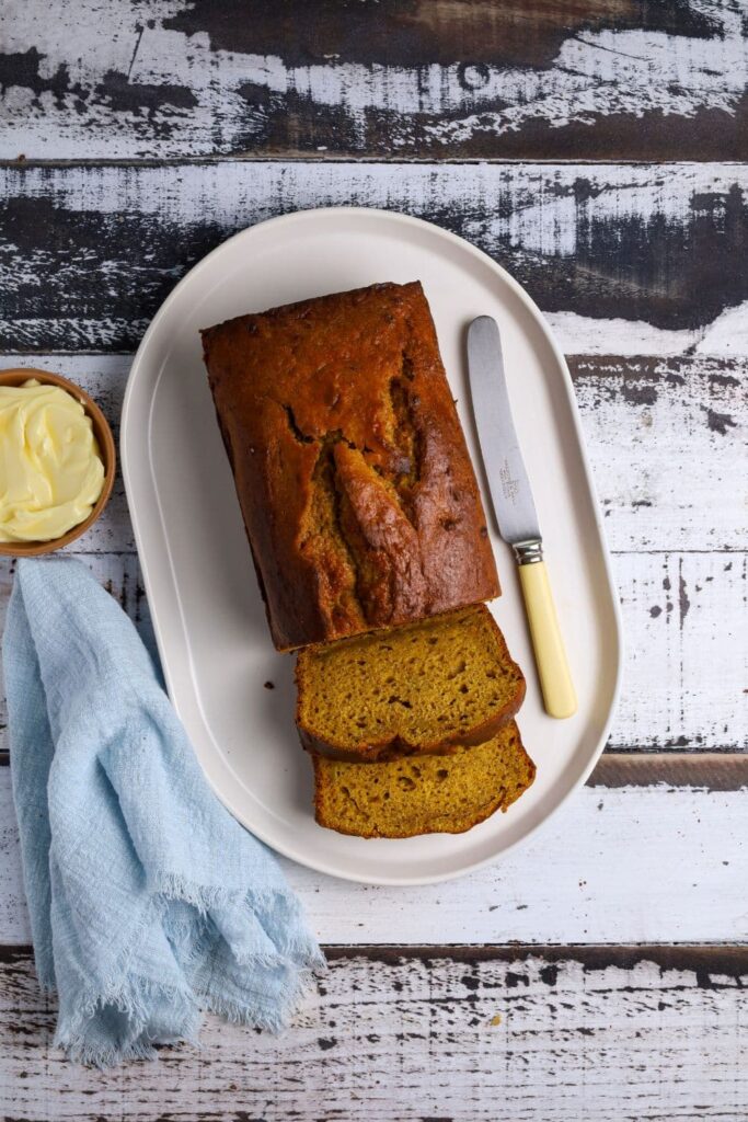 A loaf of pumpkin sourdough banana bread sitting on a white oval plate. A few slices have been sliced off the front of the loaf and there is a pale blue napkin sitting to the side of the plate.