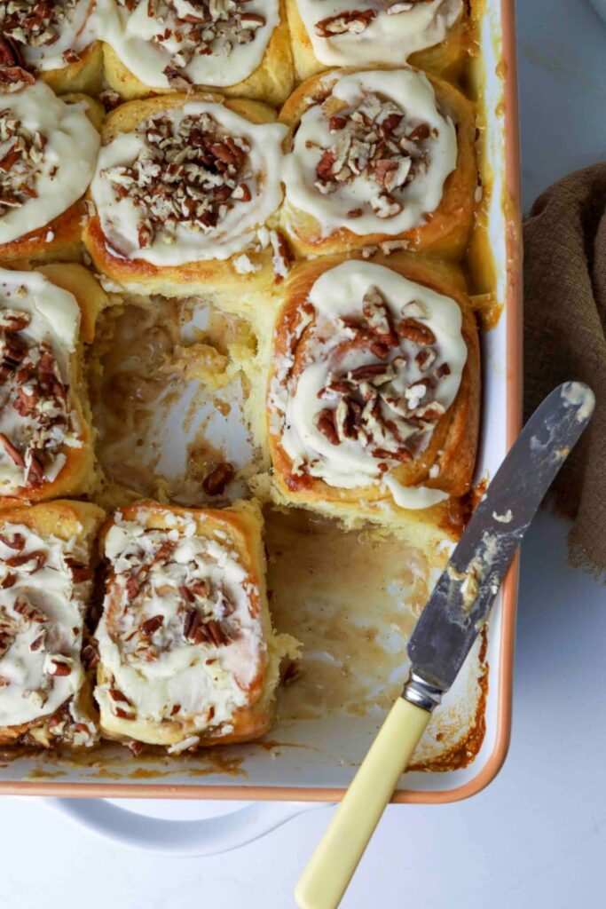 Sourdough maple pecan rolls fresh baked in baking dish with glaze on top and a knife on dish that was used to cut out two of the rolls.
