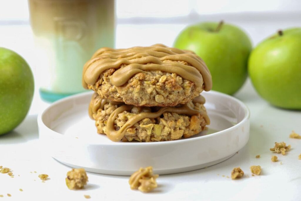 Close up of two apple cinnamon sourdough oatmeal cookies stacked on a white plate with apples in the background.