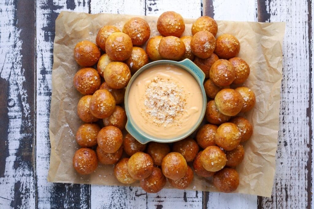 A horiztonal image showing a small green bowl filled with beer cheese dip that has been garnished with cayenne pepper. The bowl is surrounded by sourdough discard pretzel bites.