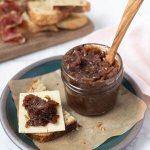 Jar of caramelized onion relish on a plate next to a piece of sourdough bread with cheese and onion relish on top.
