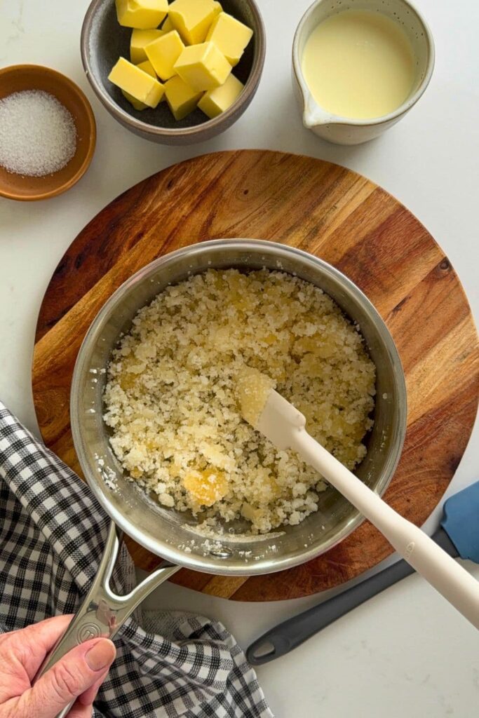 A saucepan filled with granulated sugar that is being melted. The sugar has started to melt and has formed clumps that are starting to gain some color. You can see a jug of cream, butter and dish of salt in the photo as well.