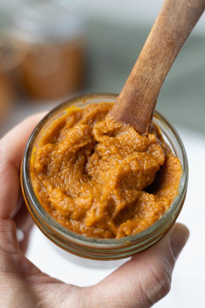 A jar of pumpkin butter being held by a hand. There is a wooden spoon in the top of the jar.