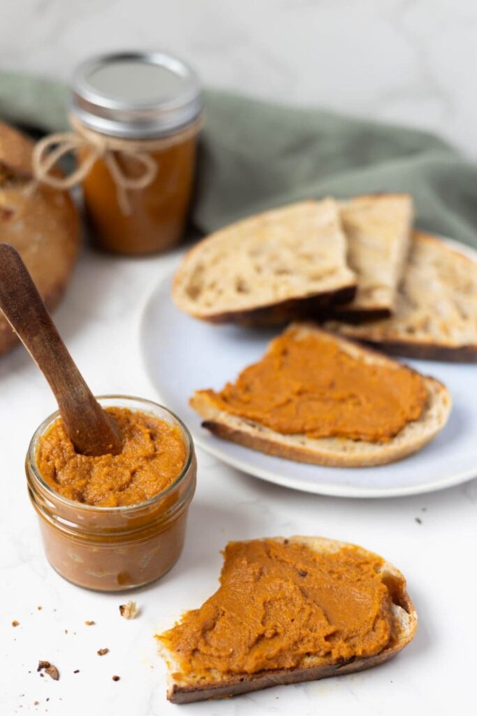 A jar of pumpkin butter with a wooden spoon inside. The pumpkin butter has been spread onto slices of sourdough bread and displayed on a white counter top in front of a sage green dish towel.