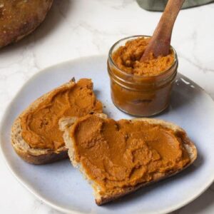 2 slices of sourdough bread spread with pumpkin butter. The jar of pumpkin butter is sitting on the plate as well, with the wooden spoon in the jar.